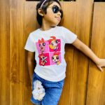 Girl wearing sunglasses and a pink headband, in a white T‑shirt with Powerpuff Girls graphics, leaning against a wooden wall.