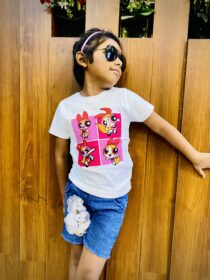 Girl wearing sunglasses and a pink headband, in a white T‑shirt with Powerpuff Girls graphics, leaning against a wooden wall.